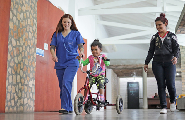 Instalaciones Teleton, niña andando en bicicleta asistida por enfermera y familiar